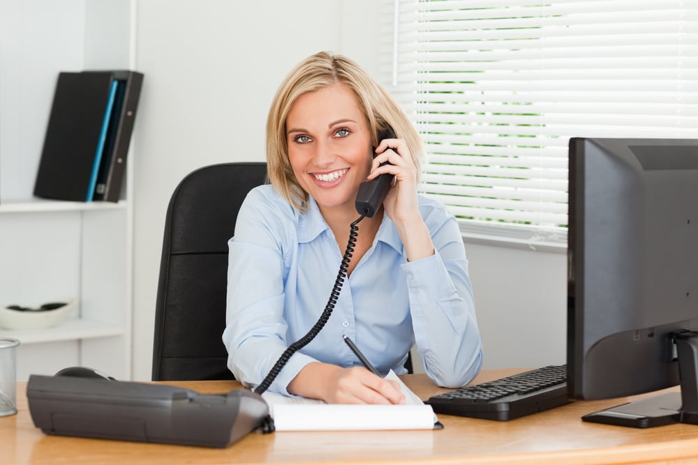 Cute businesswoman on phone writing something down looks into camera in her office